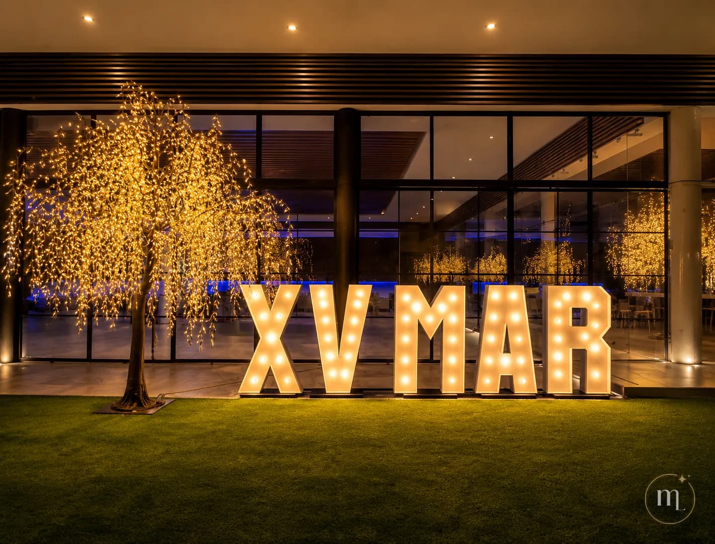 XV MAR en letras iluminadas sobre el pasto, junto a un sauce de luces, frente a un salón acristalado